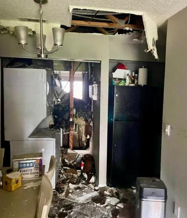 A room with severe structural damage showing an exposed ceiling with broken drywall. Debris is scattered across the room. A washing machine and dryer unit is visible amidst the damage, and a damaged electrical panel is on the right wall. A chandelier hangs from the ceiling. A room with severe structural damage showing an exposed ceiling with broken drywall. Debris is scattered across the room. A washing machine and dryer unit is visible amidst the damage, and a damaged electrical panel is on the right wall. A chandelier hangs from the ceiling.