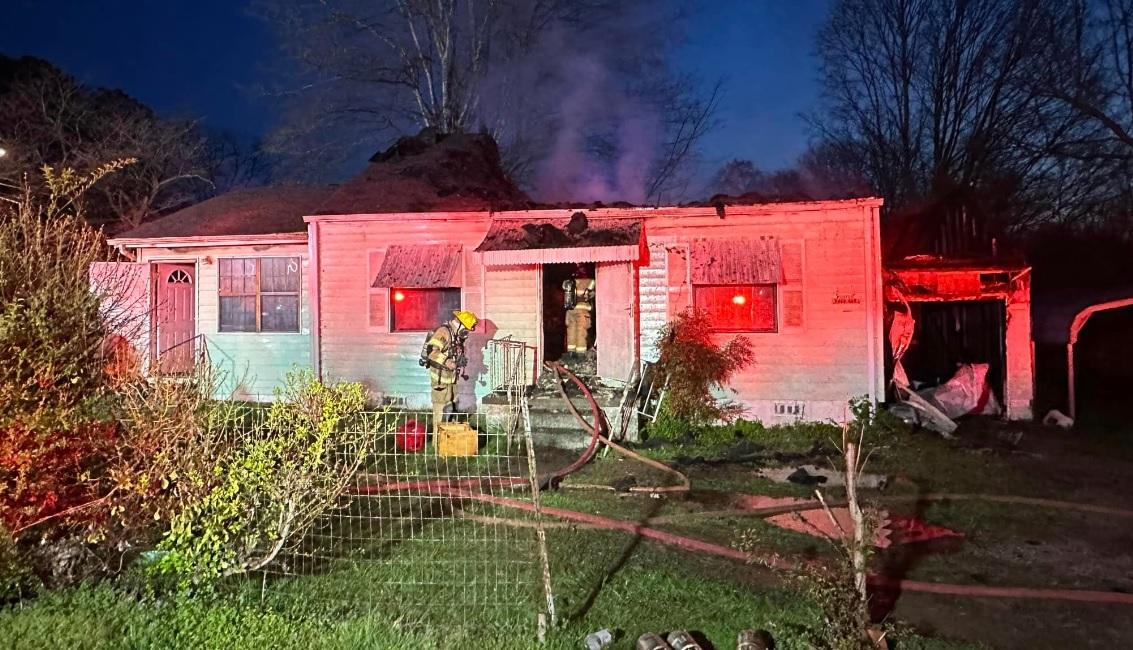 A firefighter in gear stands in front of a single-story house with a burned roof, which is emitting smoke. The scene is illuminated by red emergency lights. A garden hose is on the ground, and the surrounding area is wet from firefighting efforts. It's night time. A firefighter in gear stands in front of a single-story house with a burned roof, which is emitting smoke. The scene is illuminated by red emergency lights. A garden hose is on the ground, and the surrounding area is wet from firefighting efforts. It's night time.