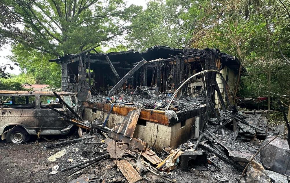 Una casa devastada por el fuego casi completamente destruida, con escombros carbonizados y madera quemada esparcidos alrededor. Los restos ennegrecidos de la estructura están rodeados de árboles altos, y un vehículo gravemente dañado está estacionado a la izquierda. La escena muestra daños y destrucción extensos.