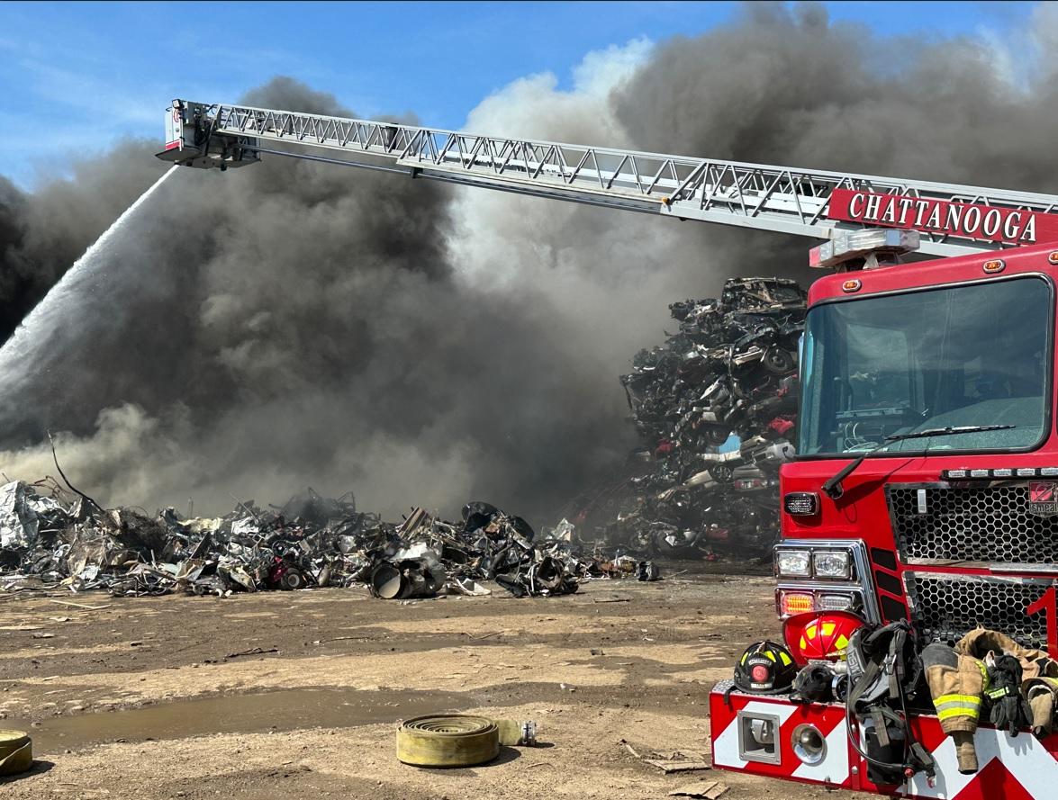 Un gran camión de bomberos con "Chattanooga" en su lateral extiende su escalera rociando agua sobre una gran pila de chatarra metálica envuelta en un denso humo negro. Hay escombros esparcidos en el suelo, y el cielo está despejado y azul al fondo.