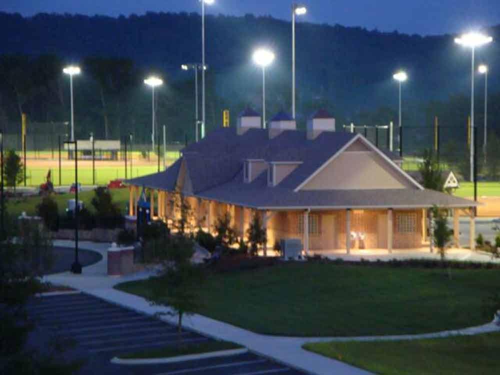 A brightly lit baseball field at night with a nearby building featuring pitched roofs and cupolas. The scene includes several light poles illuminating the field, an empty paved parking lot, and surrounding greenery. The backdrop features a dark, wooded hillside. A brightly lit baseball field at night with a nearby building featuring pitched roofs and cupolas. The scene includes several light poles illuminating the field, an empty paved parking lot, and surrounding greenery. The backdrop features a dark, wooded hillside.