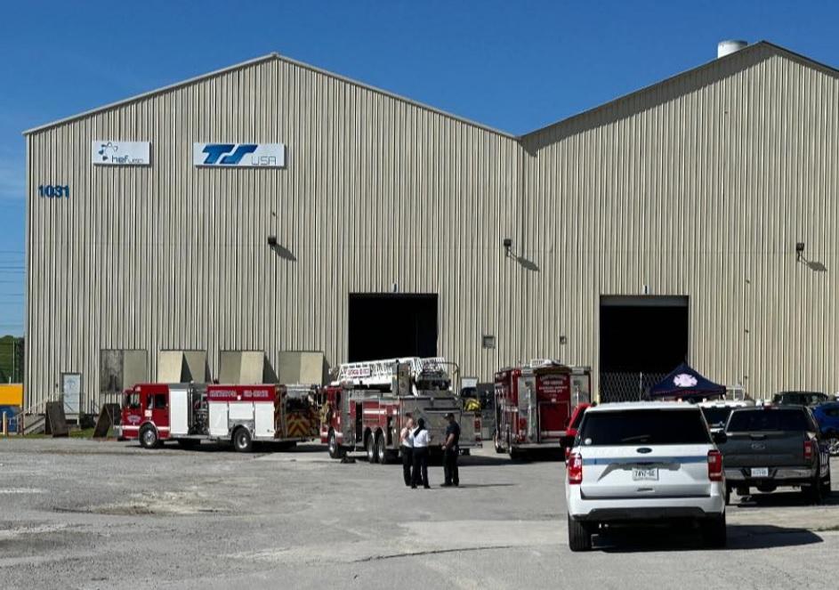 Un edificio industrial beige con el letrero "TS USA" y la dirección "1031" tiene varios vehículos de emergencia estacionados en su lote, incluidos camiones de bomberos y patrullas policiales. Bomberos y oficiales de policía están reunidos frente al edificio.