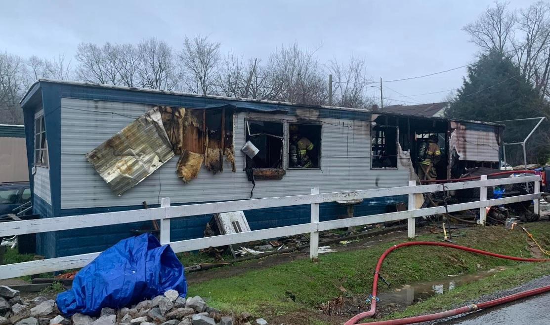 A mobile home with significant fire damage, including burnt and missing sections of the exterior, blackened windows, and charred walls. Firefighters and hoses are seen near the home, with debris scattered around. A white fence and leafless trees are visible in the background.