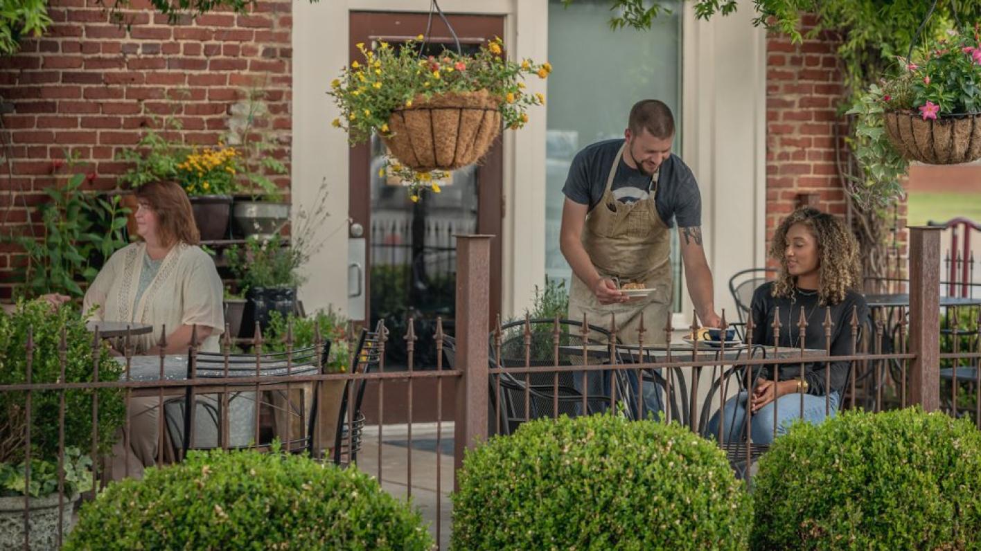 Outdoor dining scene in Chattanooga with a waiter serving a young woman while another woman sits at a nearby table, surrounded by lush greenery and hanging flower baskets.