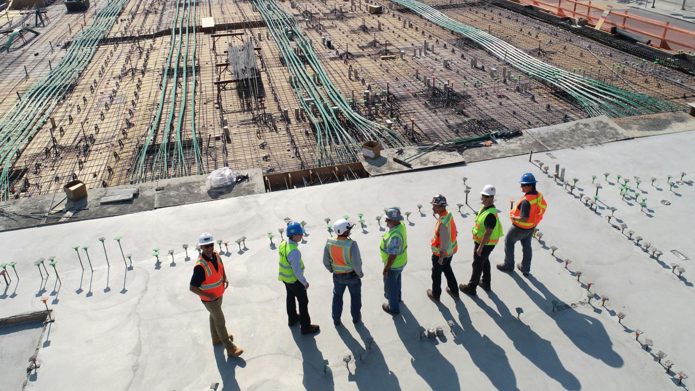 An overhead view of a construction site where a group of workers in safety gear, including hard hats and reflective vests, are gathered on a concrete slab discussing the progress. The background shows a section of the construction with exposed rebar and various construction materials.