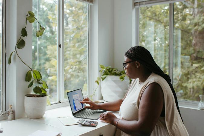 Una mujer usando una computadora portátil y una calculadora en un escritorio junto a la ventana, representando el proceso de solicitar permisos en línea.