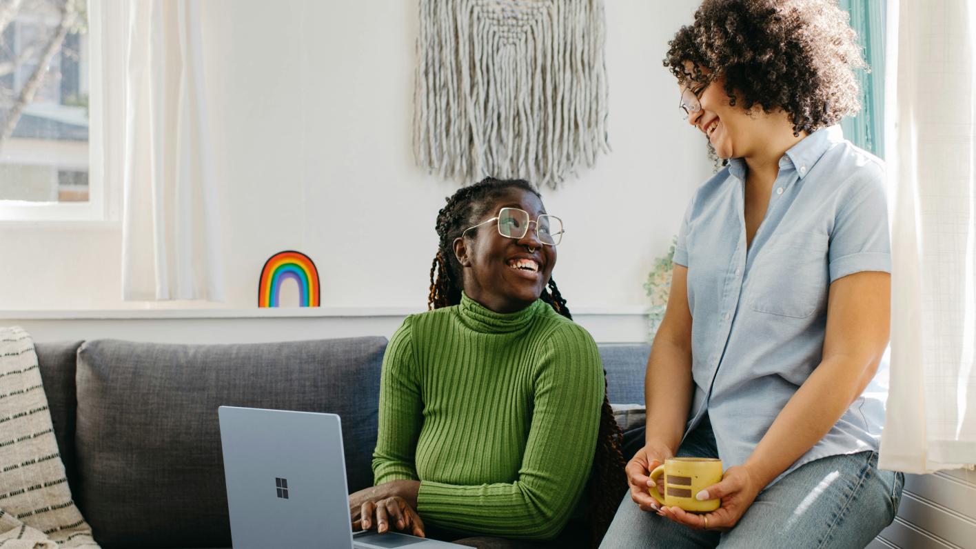 Two women smiling and conversing in a cozy living room. One is seated on a sofa with a laptop, and the other is standing, holding a coffee mug. A colorful rainbow decoration and a woven wall hanging are visible in the background.