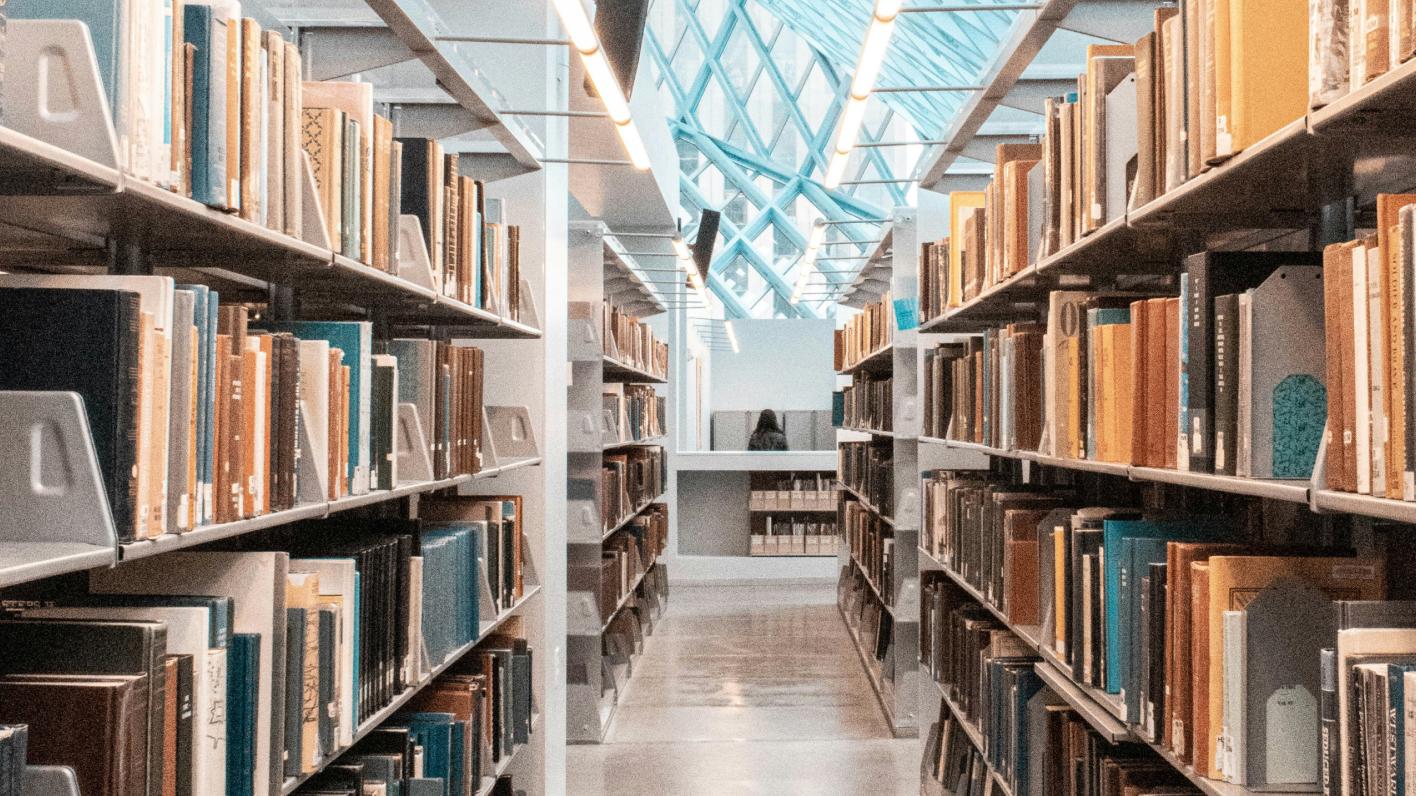 Interior view of a modern library with rows of bookshelves filled with books, leading to a bright, skylighted area at the end.