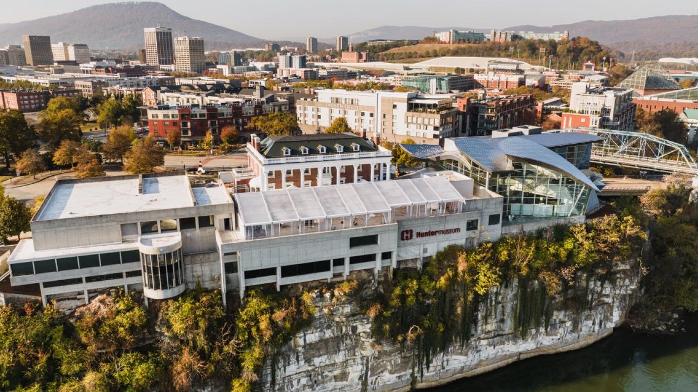 An aerial view of the Hunter Museum of American Art situated atop a cliff overlooking the Tennessee River in Chattanooga, with the city skyline and surrounding mountains in the background.