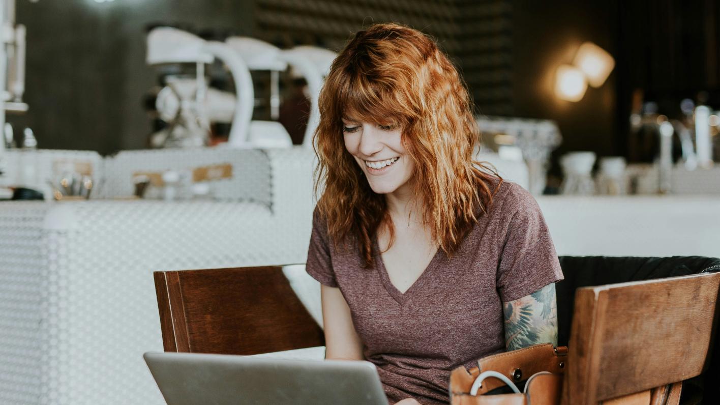 Young woman with red hair and a tattoo on her arm, smiling while using a laptop in a modern café setting