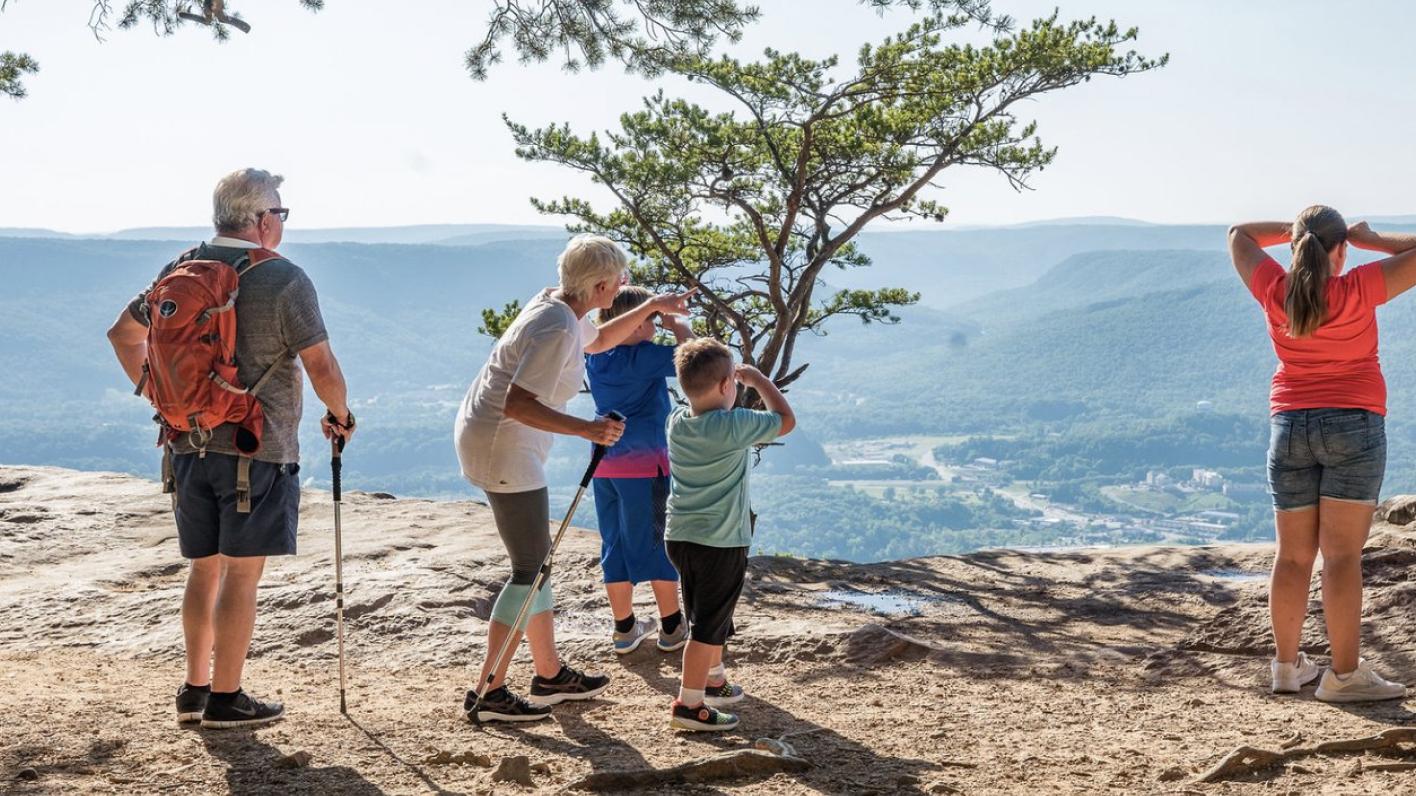 A diverse group of hikers, including children and seniors, enjoy a scenic view from a rocky overlook in Chattanooga, with lush green mountains and a distant town visible under a clear sky.